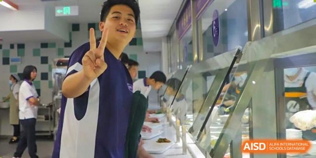 A smiling male student from Cogdel Cranleigh School Changsha in a navy shirt standing in the school's cafeteria serving line with other students.