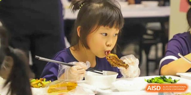 A young female student from Cogdel Cranleigh School Changsha focused on eating a piece of fried chicken or meat with chopsticks.