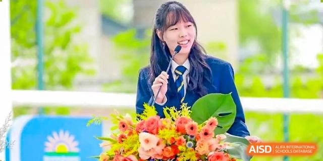 A smiling DAYSTAR ACADEMY female student in uniform speaking into a microphone, with flowers in the foreground.