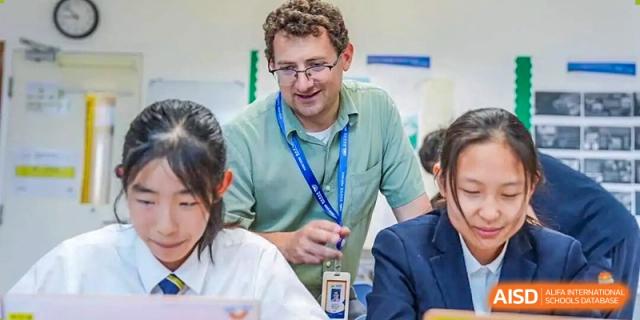 A male teacher wearing glasses and a lanyard interacts with students in a classroom setting at DAYSTAR ACADEMY.