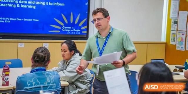 A male teacher hands out papers to students in a classroom with a screen displaying a sun graphic behind him at DAYSTAR ACADEMY.
