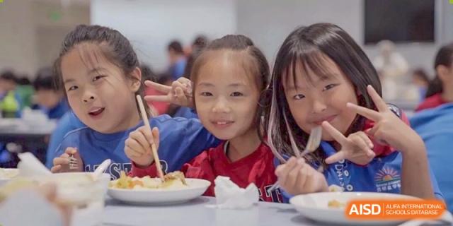 Two young female students smiling at the camera while eating a meal with chopsticks in the school cafeteria or dining hall at DAYSTAR ACADEMY.