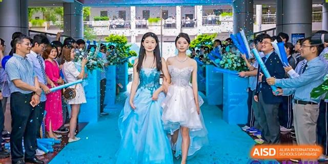 Two female students in formal light blue and white gowns walking on a blue carpet at a graduation or formal event, Fuyuan British American School.