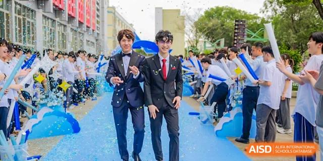 Two male students in dark suits and bow ties posing enthusiastically on a blue carpet with confetti/glitter, Fuyuan British American School.