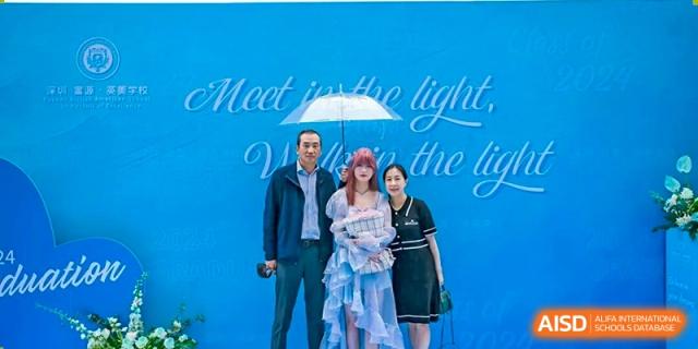 Family photo with a female graduate in a cap holding a bouquet, accompanied by two adults under an umbrella on a blue background, Fuyuan British American School.