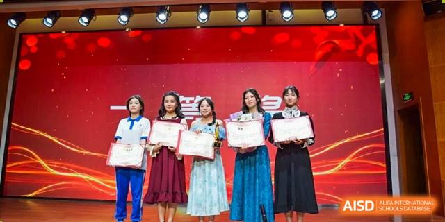 Four female students from The High School Attached to Hunan Normal University standing on a red-themed stage, holding awards or certificates after a school ceremony.