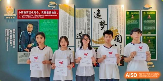Two female students and one male student holding signs with heart graphics in front of a decorative wall display at Maple Leaf International School-Zhenjiang.