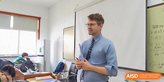 A male foreign teacher wearing a shirt and tie, holding papers, and smiling in a classroom at Maple Leaf International School-Zhenjiang.