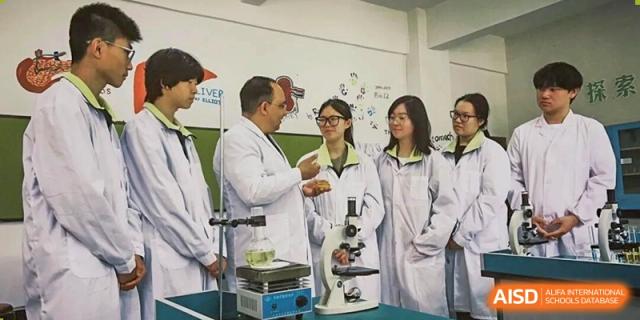 A male science teacher in a lab coat posing with two female students near a microscope in a laboratory at Maple Leaf International School-Zhenjiang.