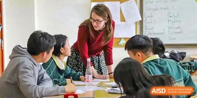 A female foreign teacher in a red top leaning in to help a small group of students at a table at Maple Leaf International School-Zhenjiang.