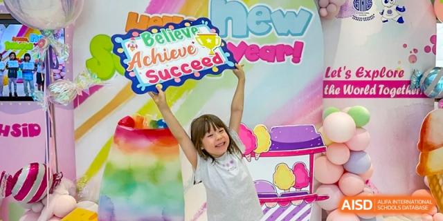 A young student smiling and holding up a colorful sign that says "Believe Achieve Succeed" at Shanghai High School International Division.