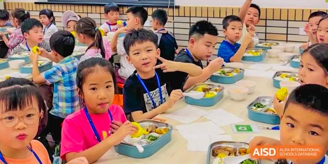 A young student smiling while eating lunch with classmates in the cafeteria or classroom at Shanghai High School International Division.