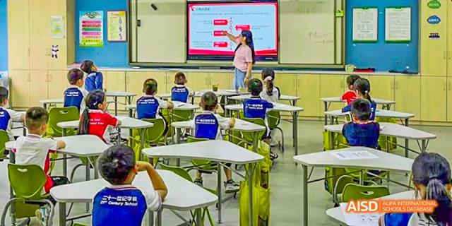 A teacher leading a lesson in a brightly lit classroom, with students seated at desks wearing their blue and white uniforms, at 21ST Century School, Wenjiang, Chengdu.