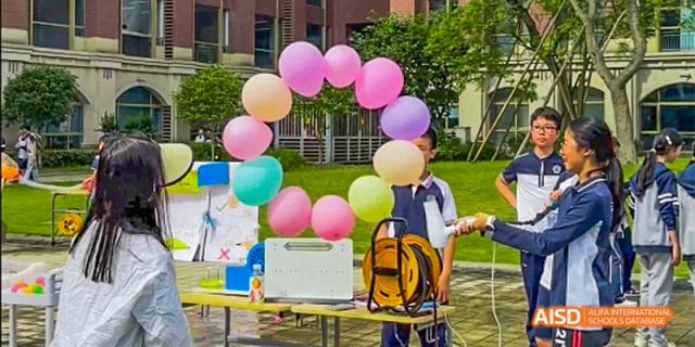 A student standing next to a table with colorful balloons and a backpack, possibly running a booth during an outdoor school fair, at 21ST Century School, Wenjiang, Chengdu.