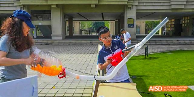 A male student conducting an outdoor science experiment with a curved pipe and a red device in the school's covered outdoor area, at 21ST Century School, Wenjiang, Chengdu.