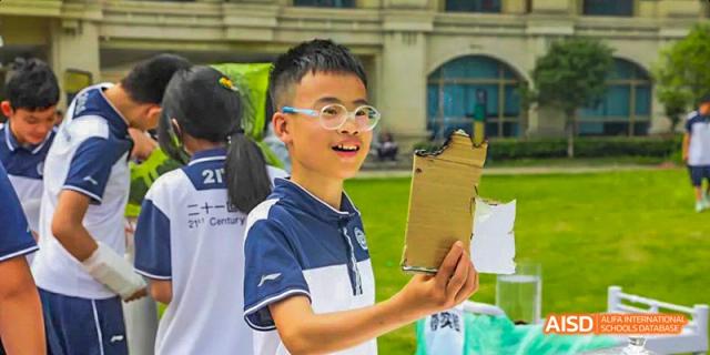 A male student in glasses holding up a piece of cardboard, looking intently at a display or during a hands-on learning activity, at 21ST Century School, Wenjiang, Chengdu.