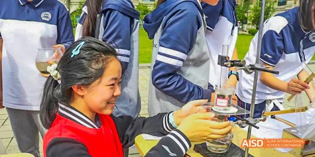 A close-up of a female student smiling while working with a small piece of scientific equipment outdoors, assisted by a teacher or peer, at 21ST Century School, Wenjiang, Chengdu.
