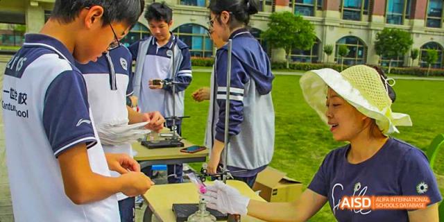 Students working on a technical project, with one person wearing gloves handling a small device on a table during an outdoor science class, at 21ST Century School, Wenjiang, Chengdu.