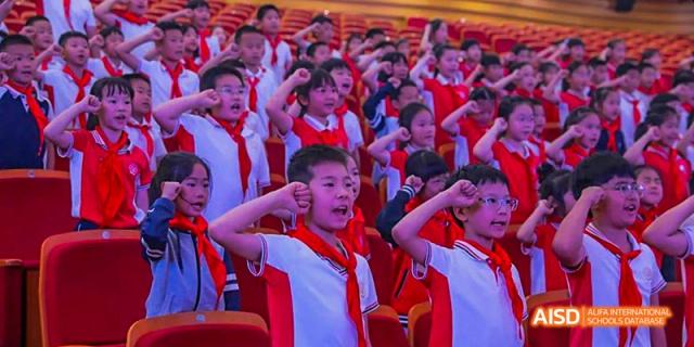 Young students in red vests cheering enthusiastically with their arms raised during a school event or performance in an auditorium, at 21ST Century School, Wenjiang, Chengdu.