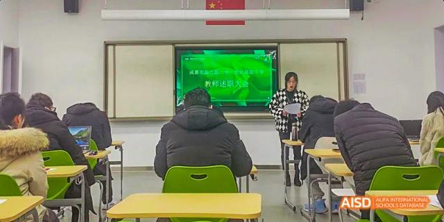A student taking a test or looking at a screen in a classroom, with another student standing nearby and a large green screen displaying Chinese text, at 21ST Century School, Wenjiang, Chengdu.