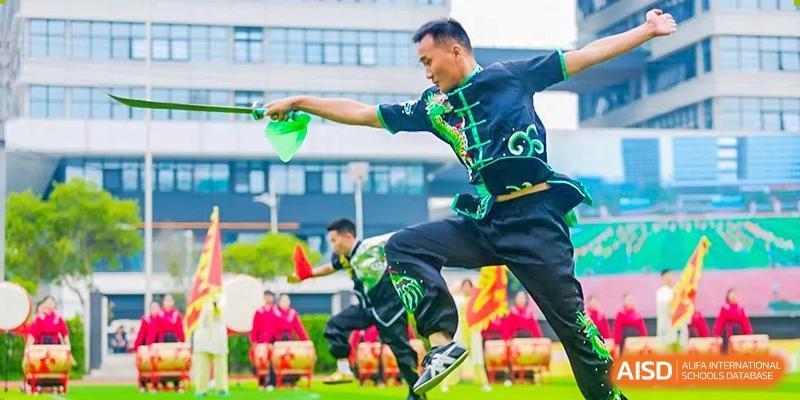 A male performer in a green and black martial arts uniform mid-action, wielding a green prop during an outdoor performance, with an audience in the background, at 21ST Century School, Wenjiang, Chengdu.