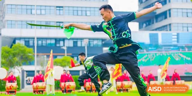 A male performer in a green and black martial arts uniform mid-action, wielding a green prop during an outdoor performance, with an audience in the background, at 21ST Century School, Wenjiang, Chengdu.