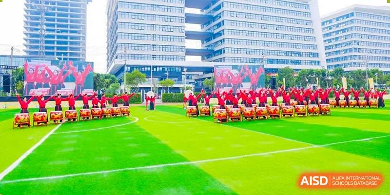 A large group performance with people in red costumes and drums on a green sports field, surrounded by modern high-rise school buildings, at 21ST Century School, Wenjiang, Chengdu.
