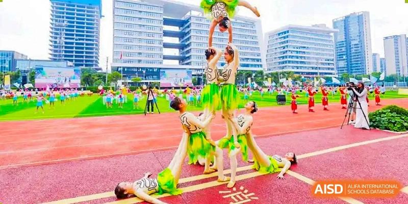 A group of students performing an acrobatic human pyramid on a track field, wearing green and yellow performance costumes, with tall school buildings in the background, at 21ST Century School, Wenjiang, Chengdu.