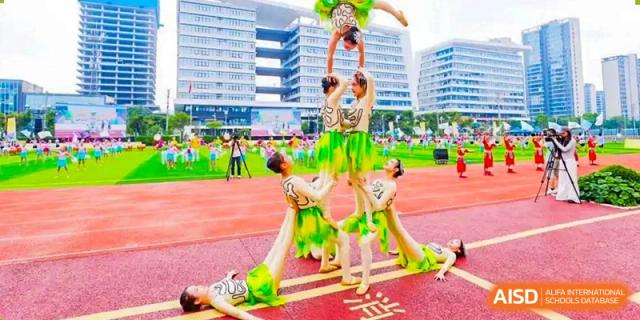 A group of students performing an acrobatic human pyramid on a track field, wearing green and yellow performance costumes, with tall school buildings in the background, at 21ST Century School, Wenjiang, Chengdu.