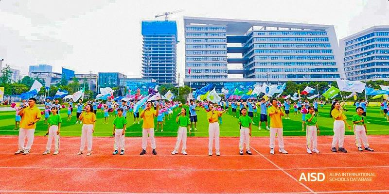 Students in yellow and green T-shirts standing on a red sports track in front of an audience and tall modern school buildings, possibly for an award or opening ceremony, at 21ST Century School, Wenjiang, Chengdu.