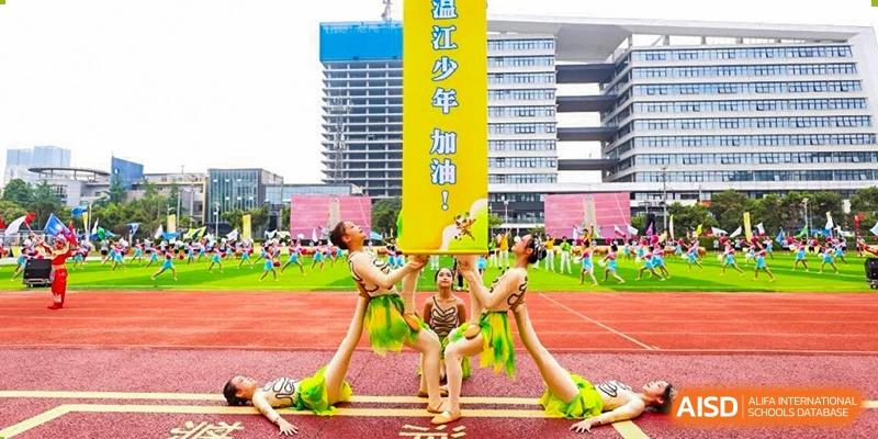 Female students performing a dance and acrobatic routine in green and yellow outfits on a sports field, with a large vertical yellow banner that has Chinese text, at 21ST Century School, Wenjiang, Chengdu.