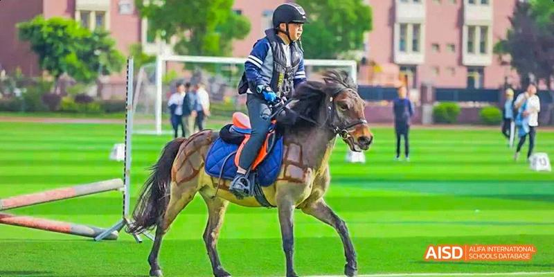 A student wearing a helmet and safety vest riding a dark brown horse across a field during an equestrian activity, with school staff and buildings in the distance, at 21ST Century School, Wenjiang, Chengdu.