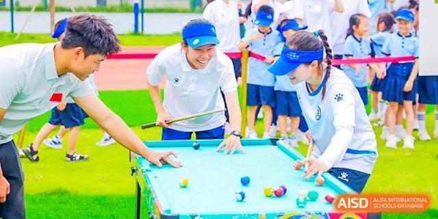Students playing billiards on an outdoor miniature pool table during a school event, supervised by an adult, at 21ST Century School, Wenjiang, Chengdu.