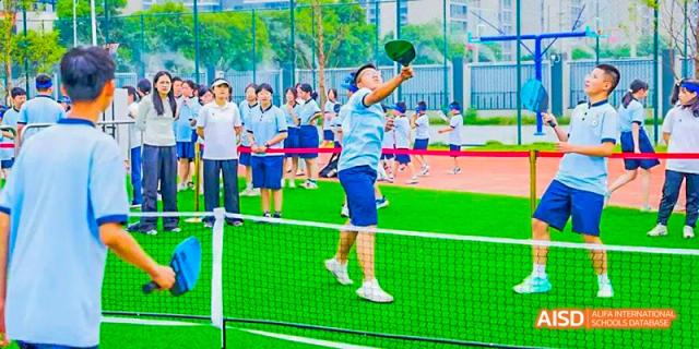 A male student jumping high to hit a ball with a table tennis paddle over a net on an outdoor court, surrounded by his classmates, at 21ST Century School, Wenjiang, Chengdu.
