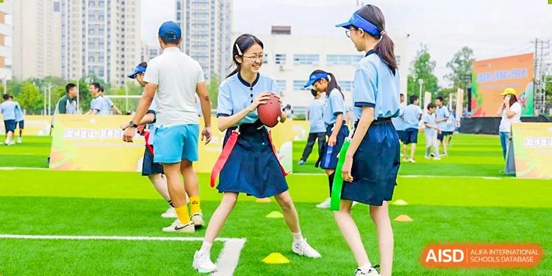A female student in a skirt and shirt holding a football, ready to play, while surrounded by other students on the sports field, at 21ST Century School, Wenjiang, Chengdu.
