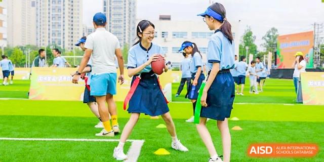 A female student in a skirt and shirt holding a football, ready to play, while surrounded by other students on the sports field, at 21ST Century School, Wenjiang, Chengdu.