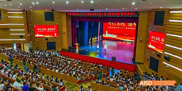 An indoor school assembly or ceremony in a large auditorium, with a speaker on stage and a large audience of students seated in the stands, at 21ST Century School, Wenjiang, Chengdu.