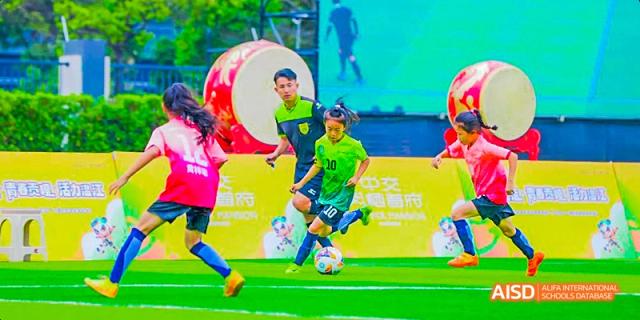 Two students playing football (soccer) on a turf field, wearing matching green jerseys, with a large drum and a banner in the background, at 21ST Century School, Wenjiang, Chengdu.