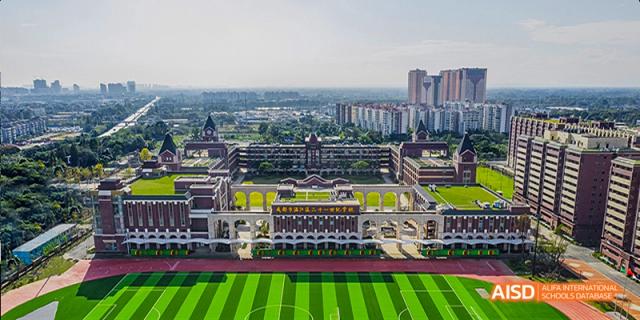 An aerial view of the expansive school campus, showing the main academic buildings and a large green sports field, surrounded by a cityscape, at 21ST Century School, Wenjiang, Chengdu.
