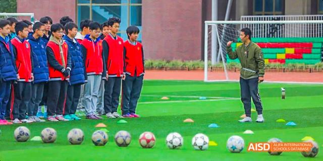 A line of students in red jackets standing on a green sports field with several soccer balls and training cones lined up in front of them, at 21ST Century School, Wenjiang, Chengdu.
