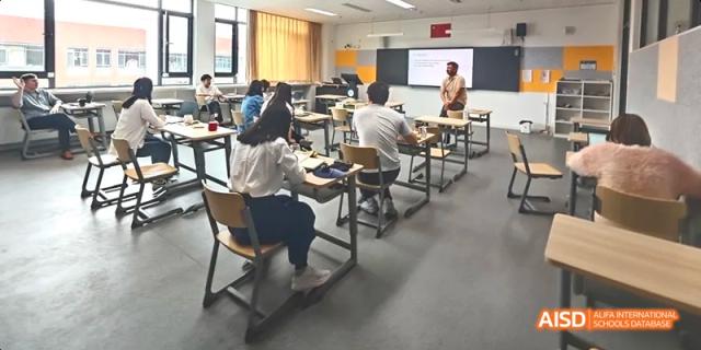 Students taking notes while seated at desks in a classroom with a teacher standing near a large screen at BDA School of The High School Affiliated to Renmin University of China.
