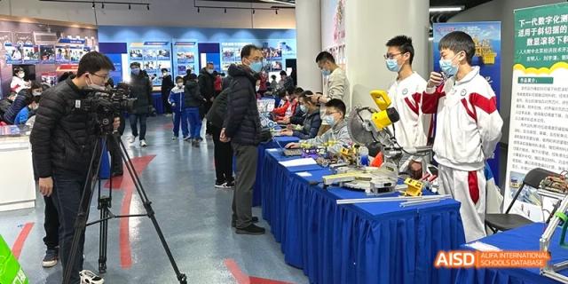 Students and adults gathered around tables viewing various electronic or robot projects displayed at a school fair or exhibition at BDA School of The High School Affiliated to Renmin University of China.