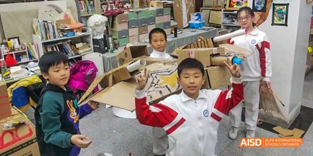 A young student in a red and white uniform holding up a hand next to their cardboard creation at BDA School of The High School Affiliated to Renmin University of China.