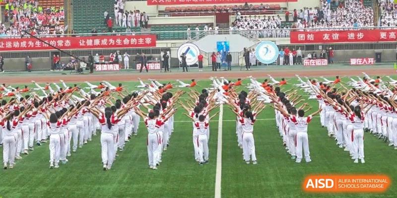 Students performing a synchronized routine on a green sports field in front of a large crowd in the stands at BDA School of The High School Affiliated to Renmin University of China.