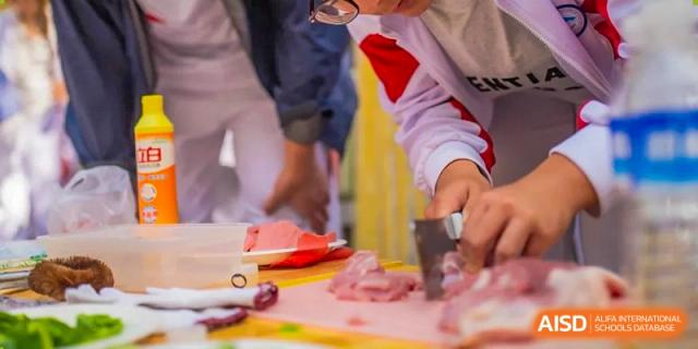 Close-up of a student from BDA School of The High School Affiliated to Renmin University of China using a knife to cut raw meat on a pink cutting board during a cooking or life skills event.