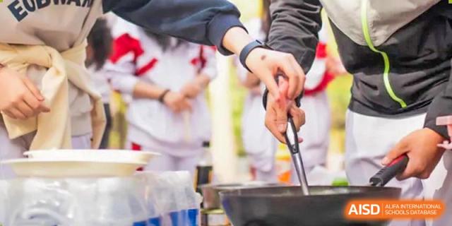 A student from BDA School of The High School Affiliated to Renmin University of China using tongs to handle food in a small pot or pan outdoors.