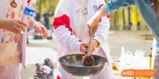 Close-up of a student from BDA School of The High School Affiliated to Renmin University of China in uniform cooking outside, stirring food with a ladle in a black wok.