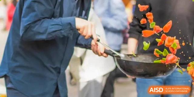 Two students from BDA School of The High School Affiliated to Renmin University of China in an outdoor setting, collaboratively cooking and holding a black wok.