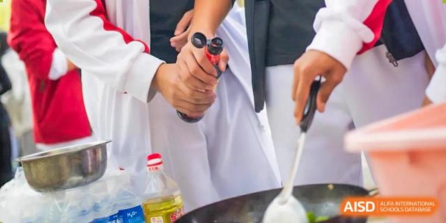 Close-up of a student in a white apron from BDA School of The High School Affiliated to Renmin University of China holding a bottle of cooking oil or sauce over a wok during a cooking activity.