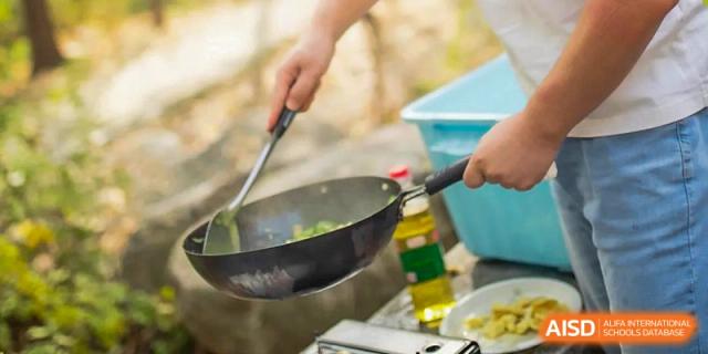 A student from BDA School of The High School Affiliated to Renmin University of China stirring green vegetables and other food in a black wok over a portable burner during an outdoor cooking class.
